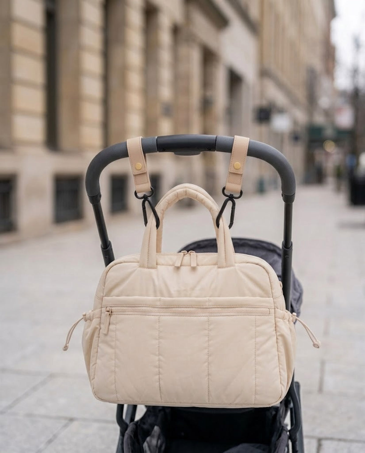 Sac crème accroché à la poignée d’une poussette par deux crochets beiges, dans une rue Image